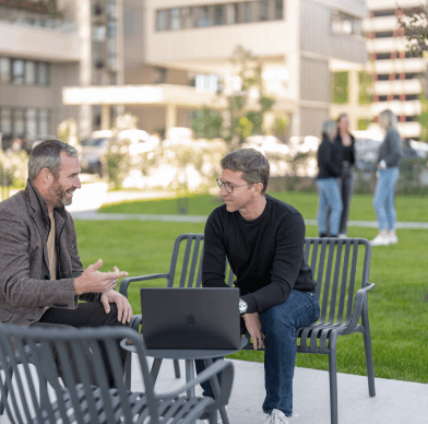 Zwei Männer sitzen im Grünen mit Laptop, moderne Gebäude im Hintergrund.