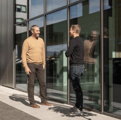 Zwei Männer unterhalten sich vor moderner Glasfassade eines Wohngebäudes, reflektiert im Fenster.