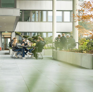Menschen sitzen an Tischen auf Terrasse moderner Wohnanlage, umgeben von Grünflächen.